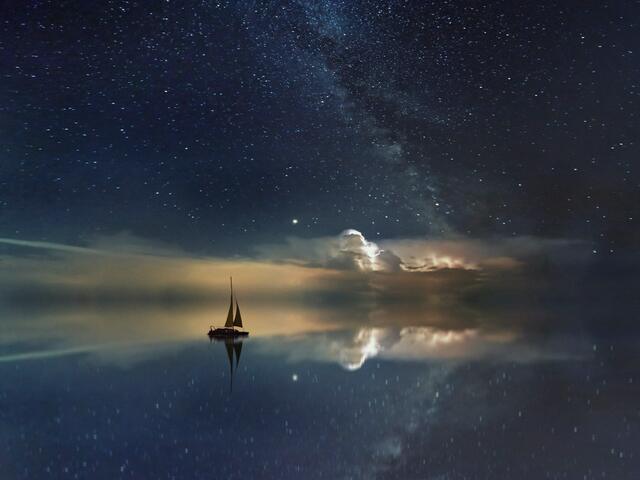 A sailing boat on calm water at night, under a starry sky.